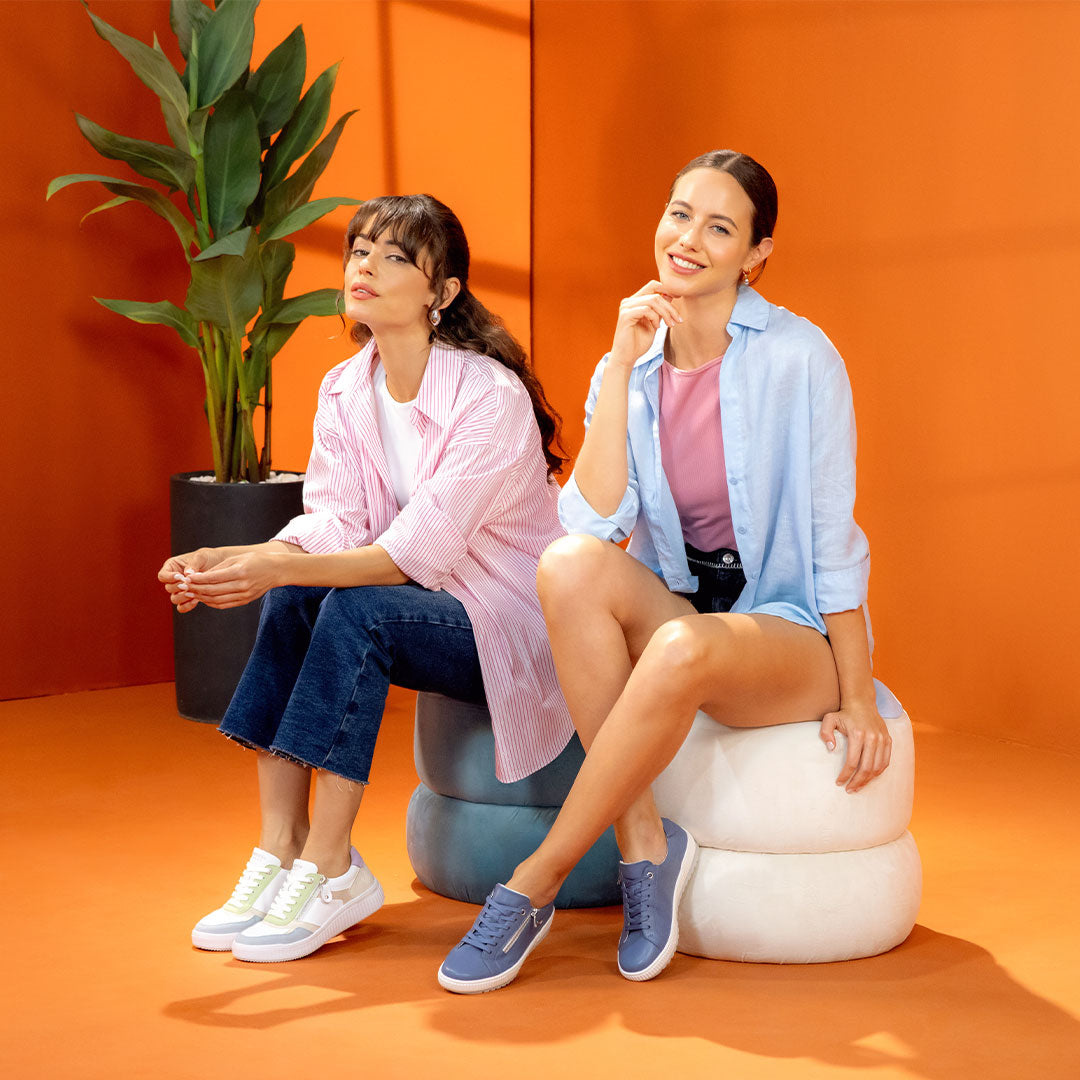 Two women wearing remonte sneakers, seated against an orange backdrop